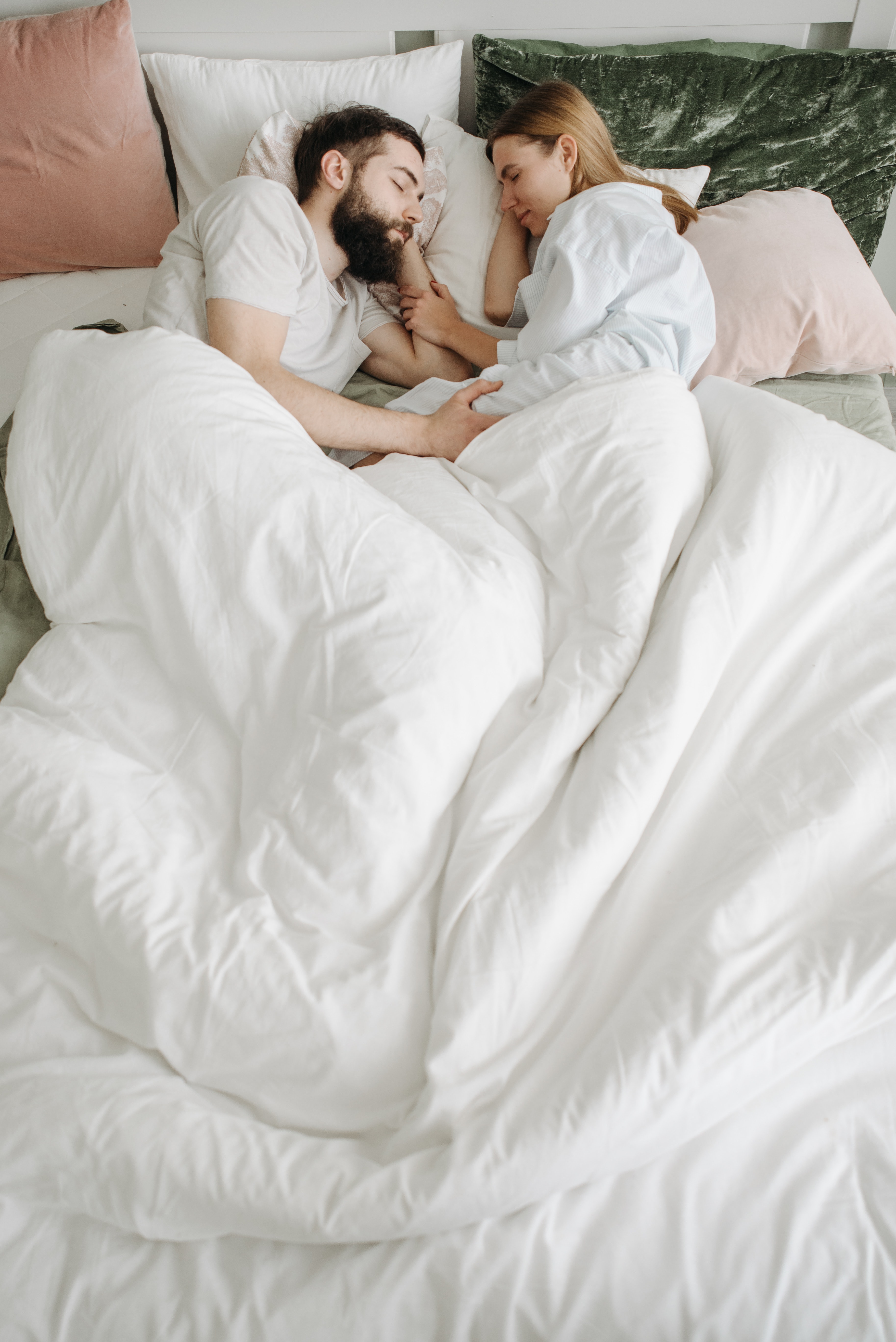 white mixed-gender couple laying in a bed with white and pink pillows and a white blanket, laying facing each other and with their legs touching under the blankets