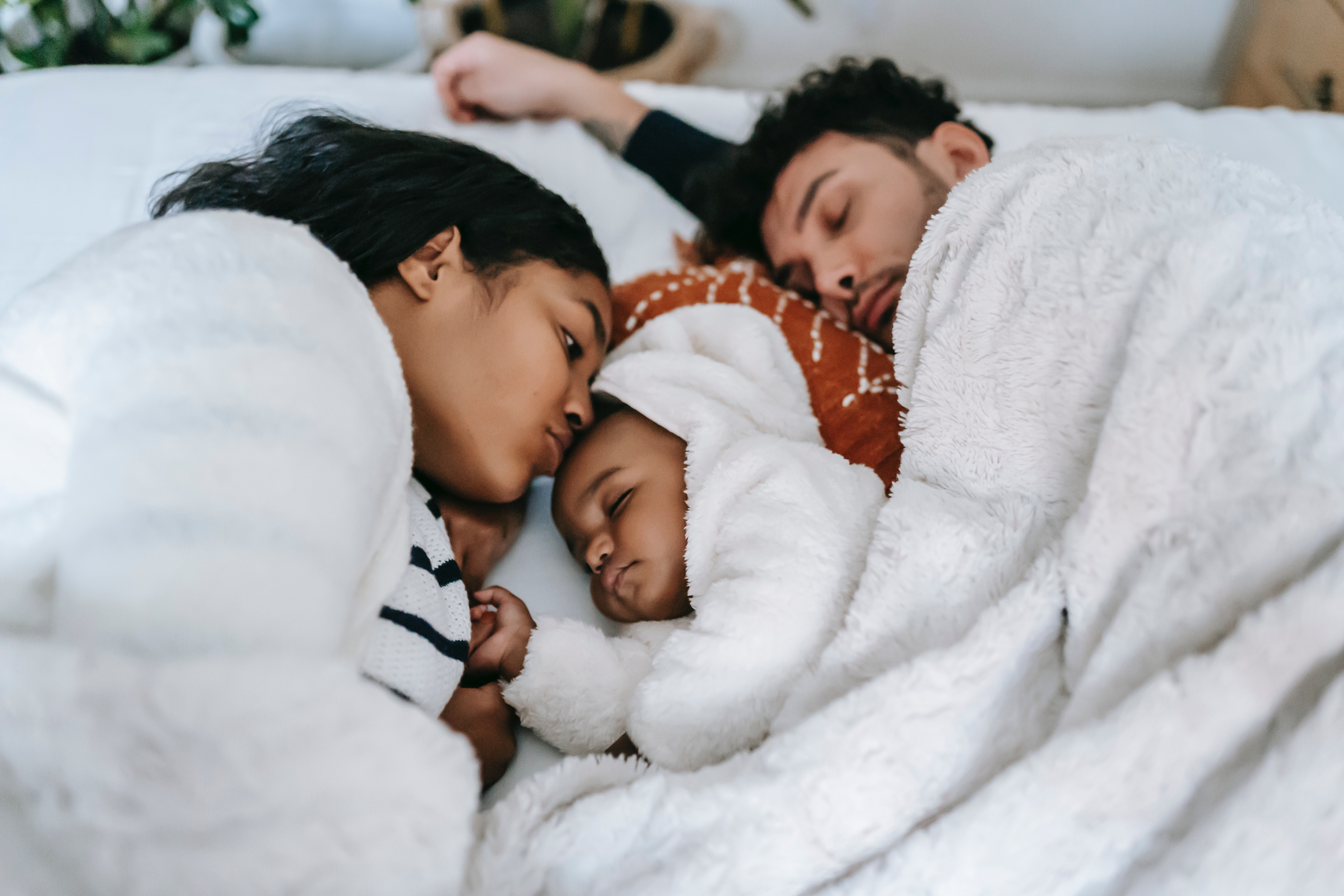 A mixed gender couple cuddles together with a baby in between them on a bed, sharing a white blanket.