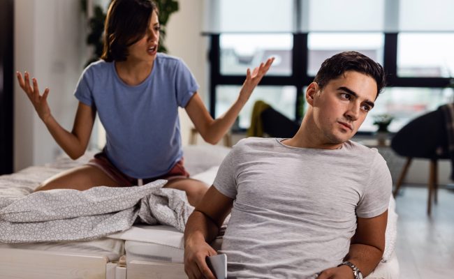 Young man sitting by the bed and ignoring his girlfriend who is arguing with him.
